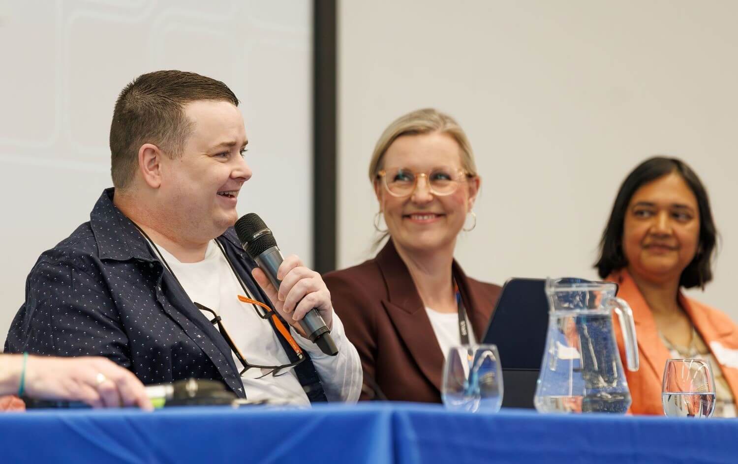 A photo of three people sitting at a table and smiling. One of them has a microphone and is speaking.